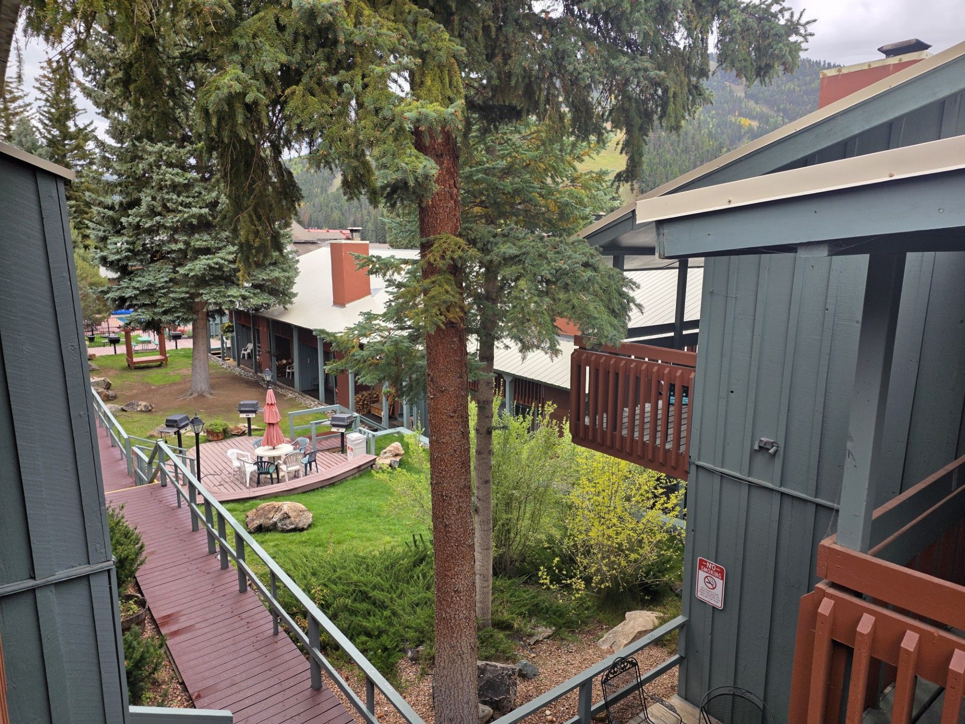 Rainy garden walkway beside townhouses, with trees, red path, and a small landscaped courtyard.