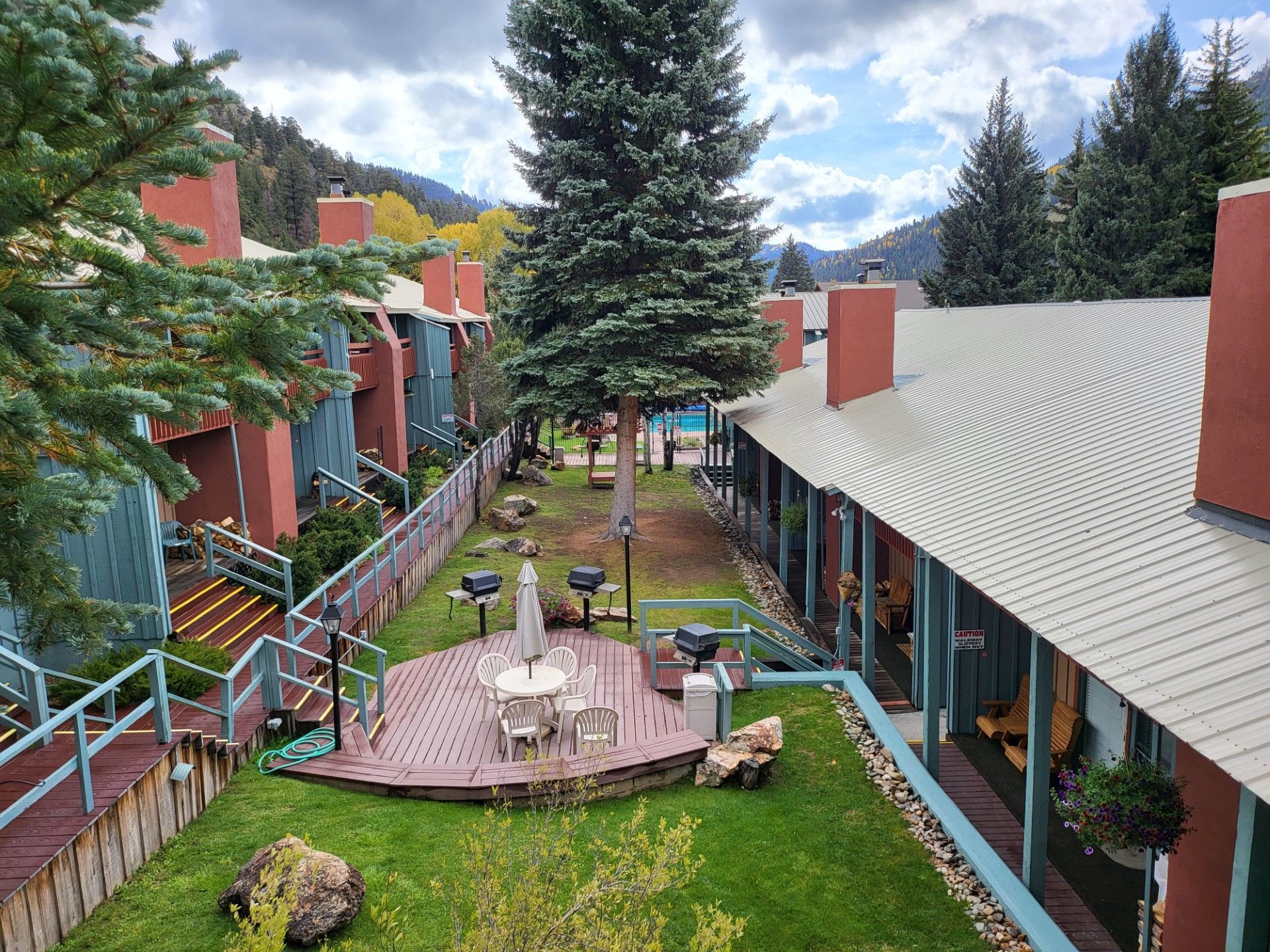 Courtyard between red buildings with a wooden deck, outdoor tables, green lawn, and tall evergreen trees
