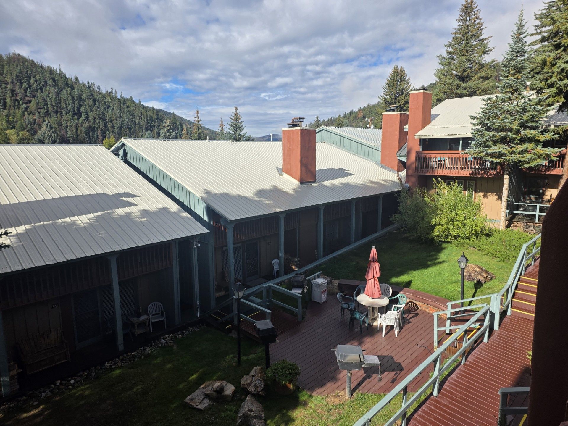 Mountain resort courtyard with patio tables, red umbrellas, green lawn, and forested hills beyond