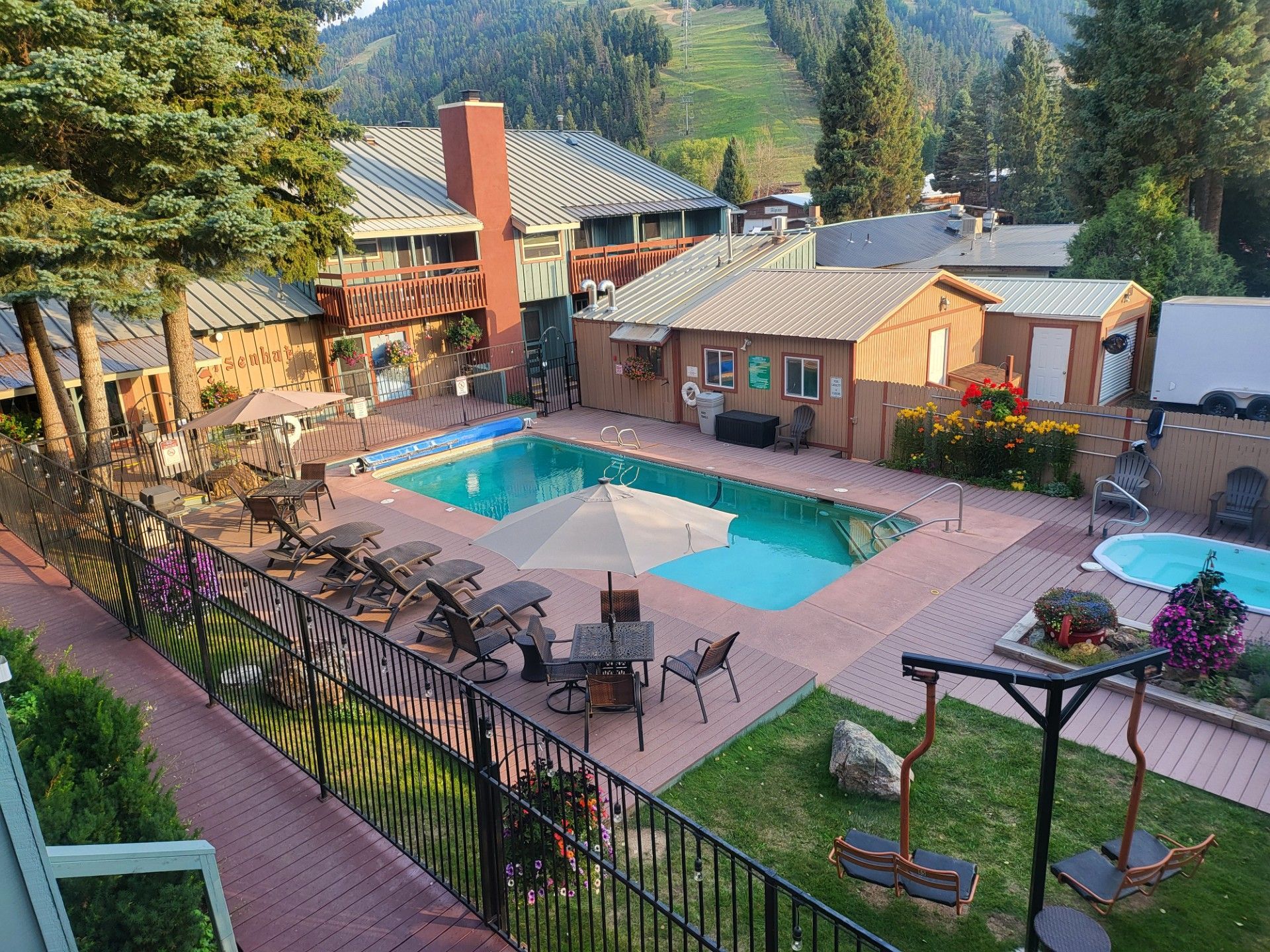 Resort-style courtyard with a blue pool, lounge chairs, patio tables, and small buildings among trees