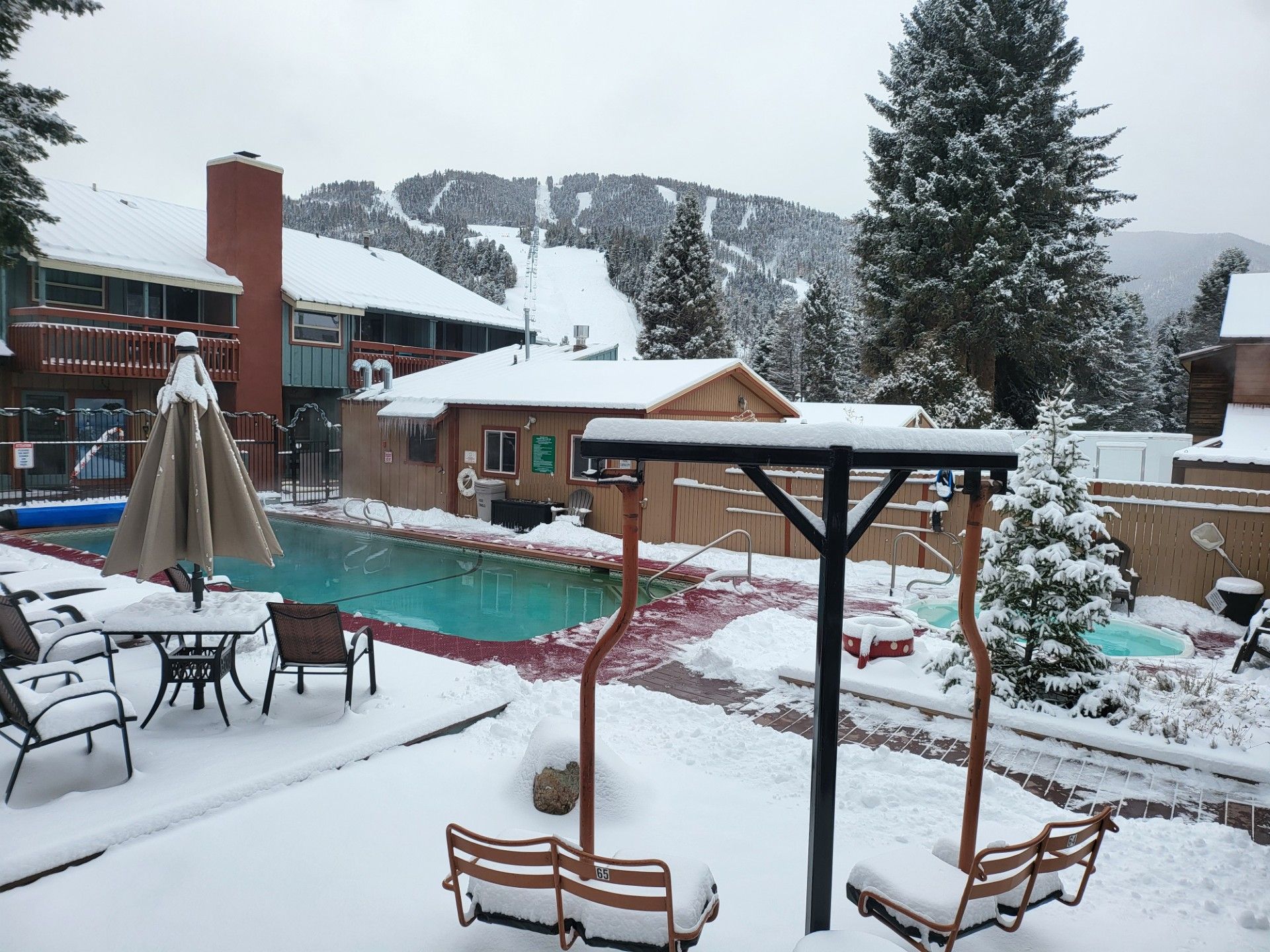 Snowy resort pool patio with lounge chairs, swings, and mountain backdrop