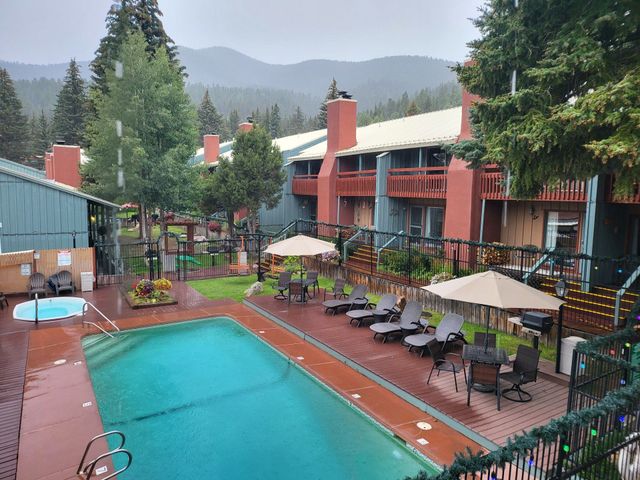 Outdoor hotel pool with lounge chairs, umbrellas, and red buildings against a mountain backdrop