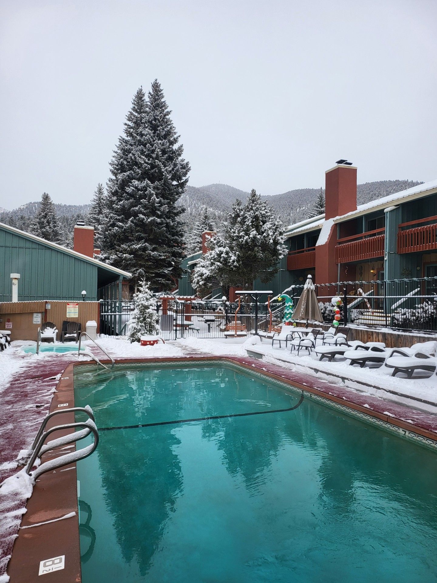 Snowy resort pool with turquoise water, lounge chairs, and evergreen trees amid mountain buildings