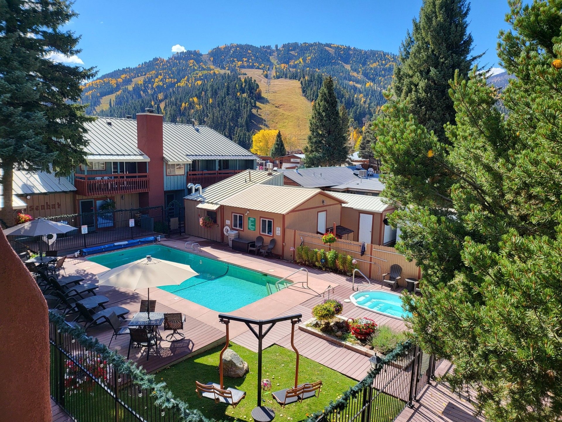 Mountain resort courtyard with turquoise pools, lounge chairs, and tall pine trees under a clear blue sky