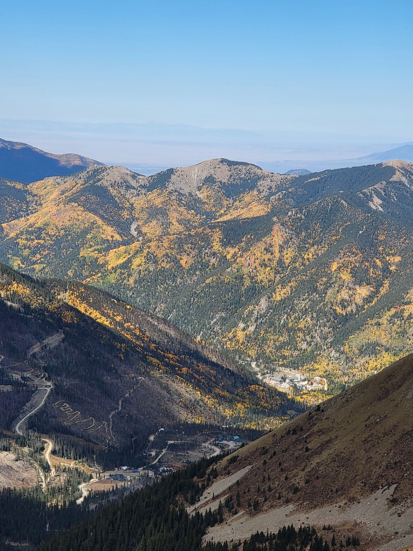 Mountain valley with sunlit autumn slopes, rugged ridges, and a winding road below under a clear blue sky