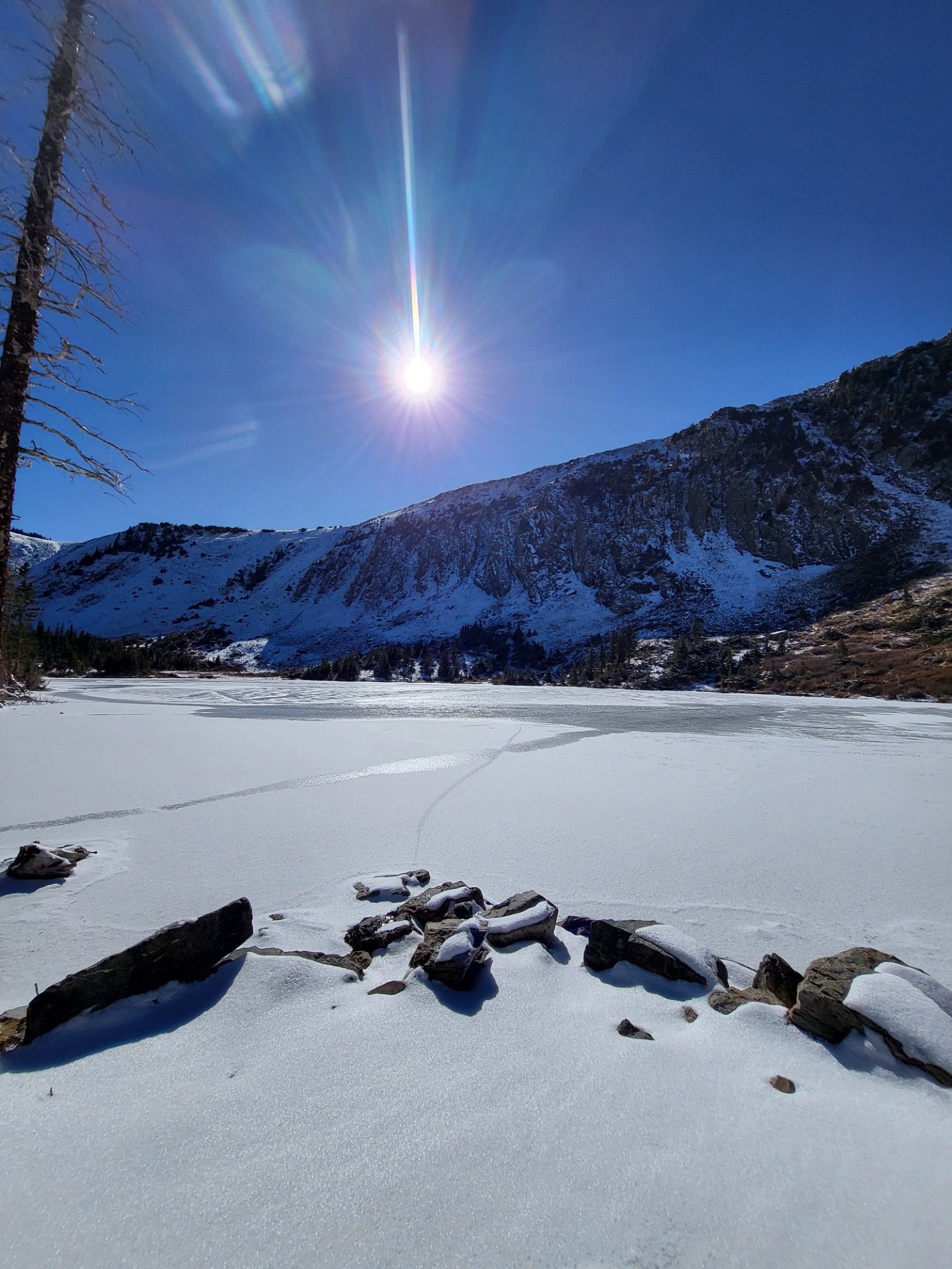 Snowy frozen lake beneath bright sun and blue sky, with mountains in the background and rocks in the foreground