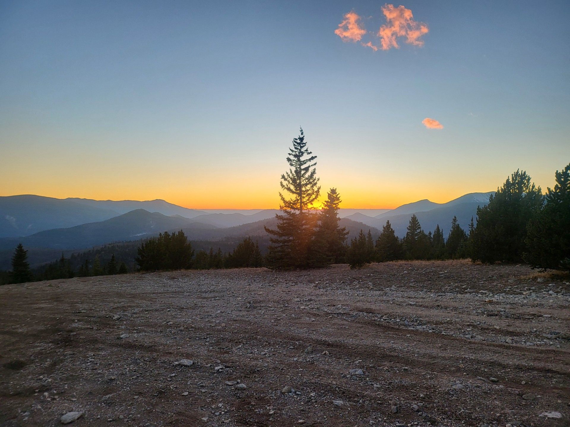 Sunset over mountain ridges with a lone pine tree and rocky foreground under a glowing sky