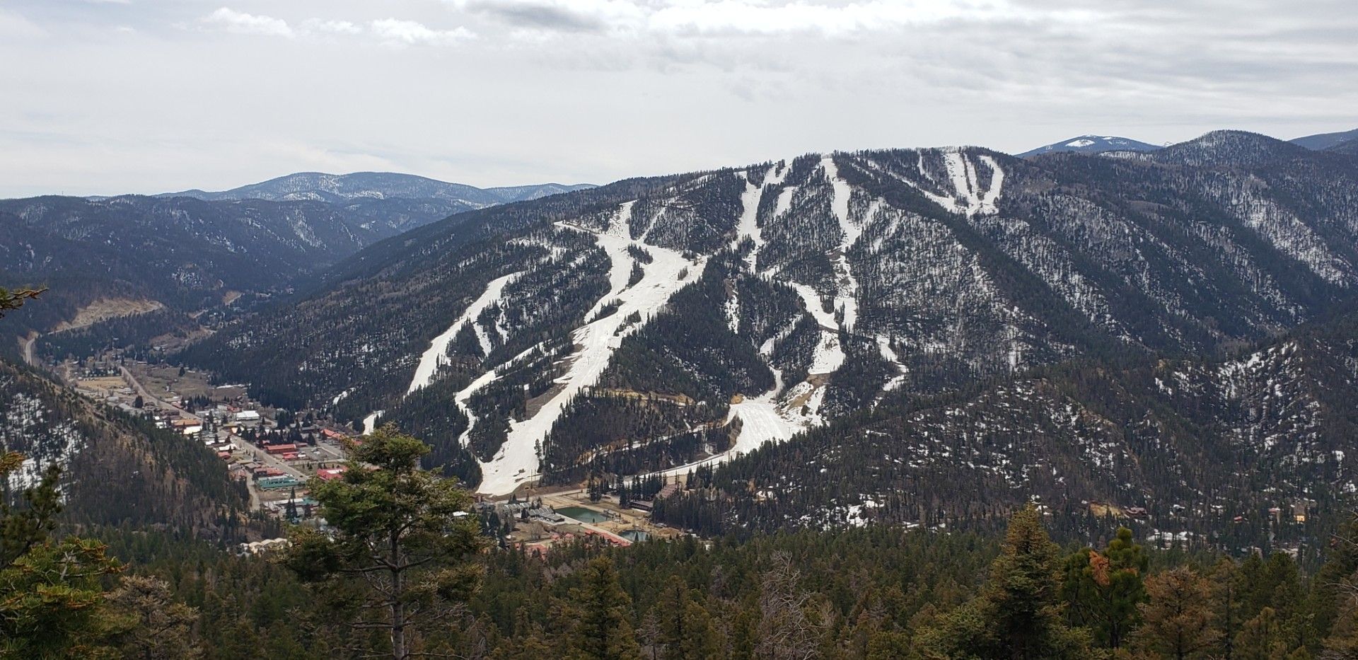 Snowy mountain ski slopes under a cloudy sky, with forested ridges in the foreground