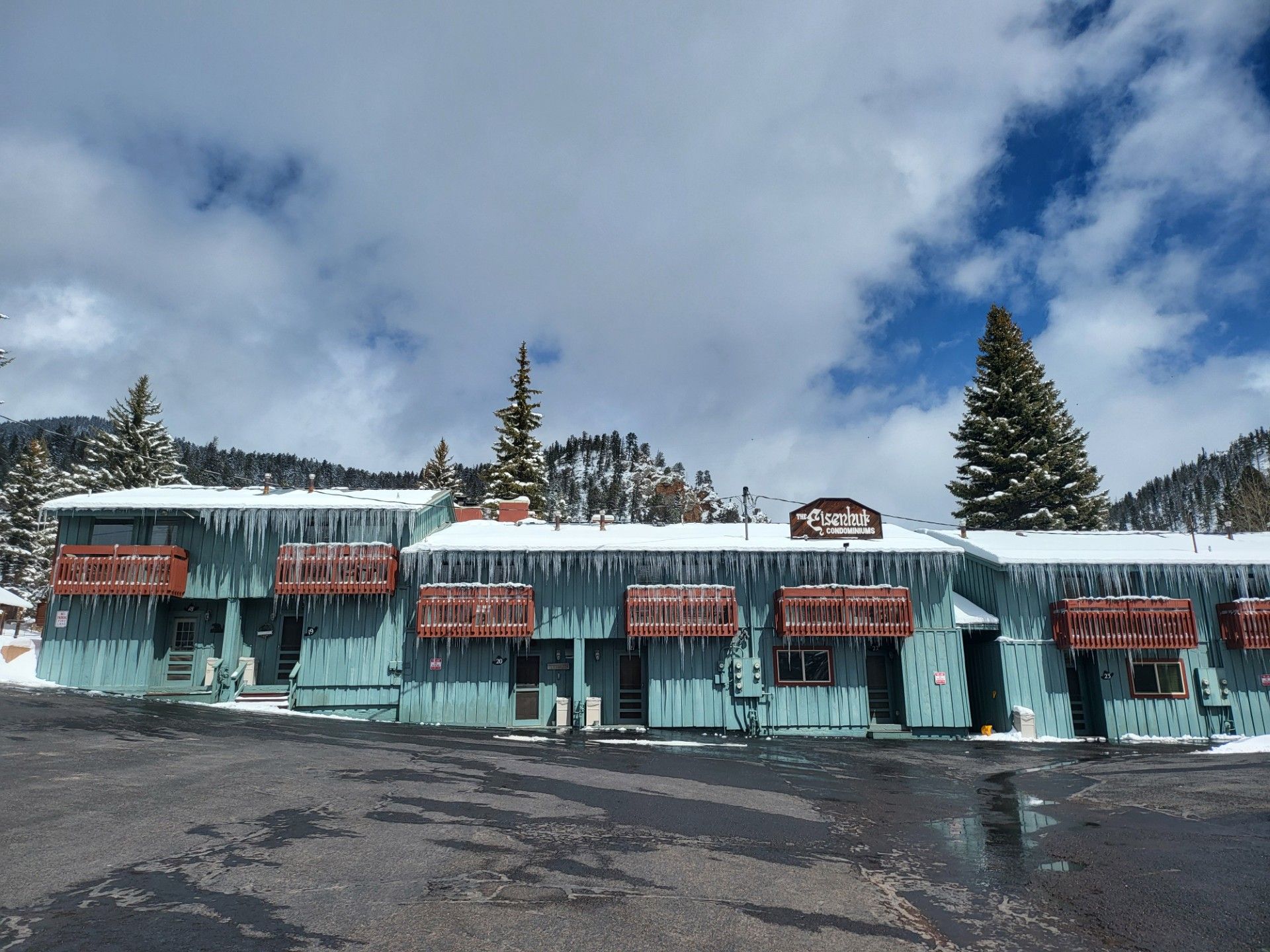 Snow-covered turquoise lodge with red awnings under a cloudy sky and tall pine trees