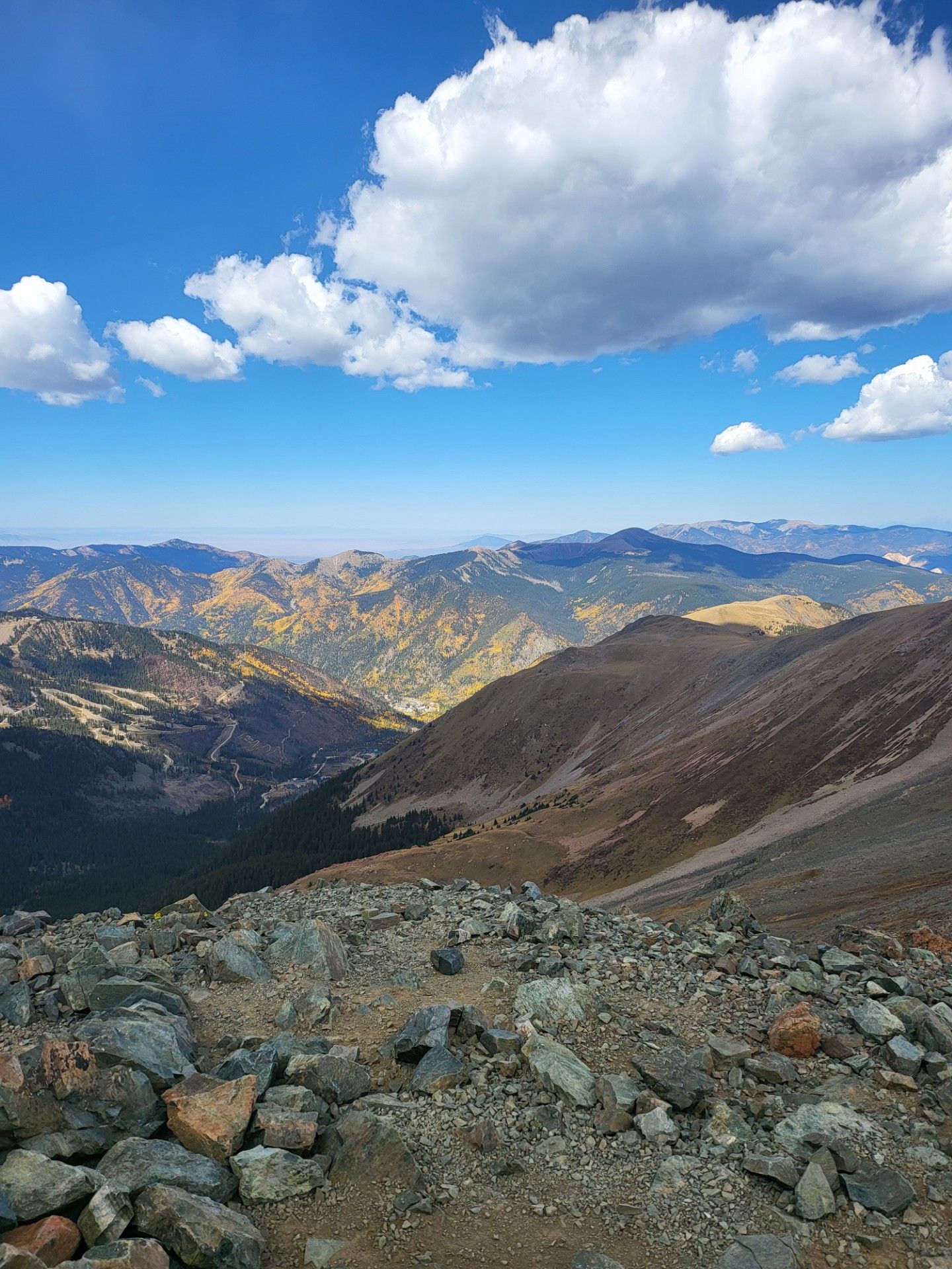 Rocky mountain overlook with distant peaks, a valley below, and a bright blue sky with clouds.
