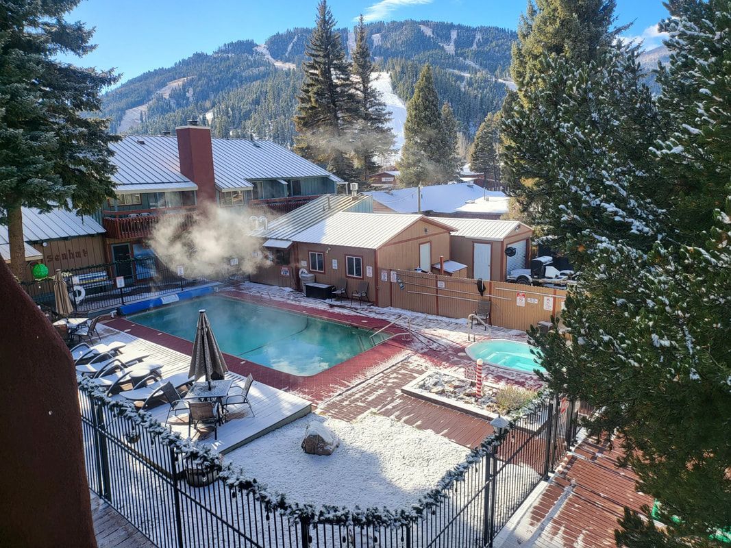 Snowy mountain resort pool with steaming hot tub, red deck, and pine trees