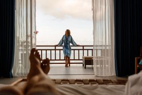 Person standing on a balcony in a blue robe, seen from a bed with sheer curtains framing the view