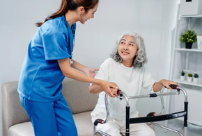 Caregiver in blue uniform helping a smiling elderly woman stand with a walker in a bright living room