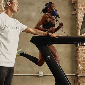 Two runners on treadmills in a gym; one wears a blue mask and the other points while walking beside her