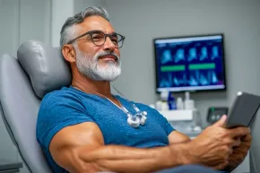 Man reclining in a clinic chair, smiling at a tablet with medical monitors in the background