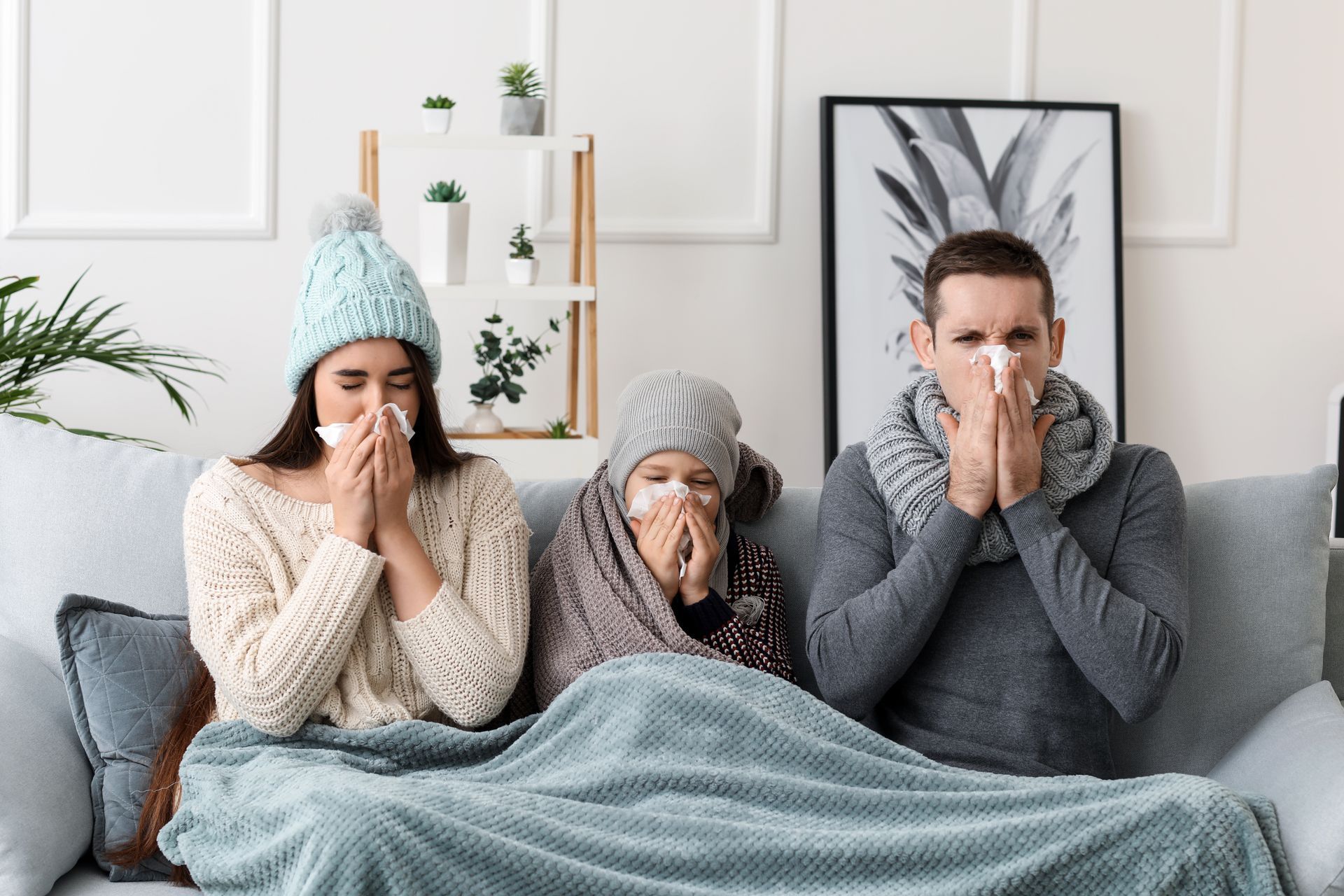 Three people on a couch bundled in blankets, holding tissues and appearing sick.