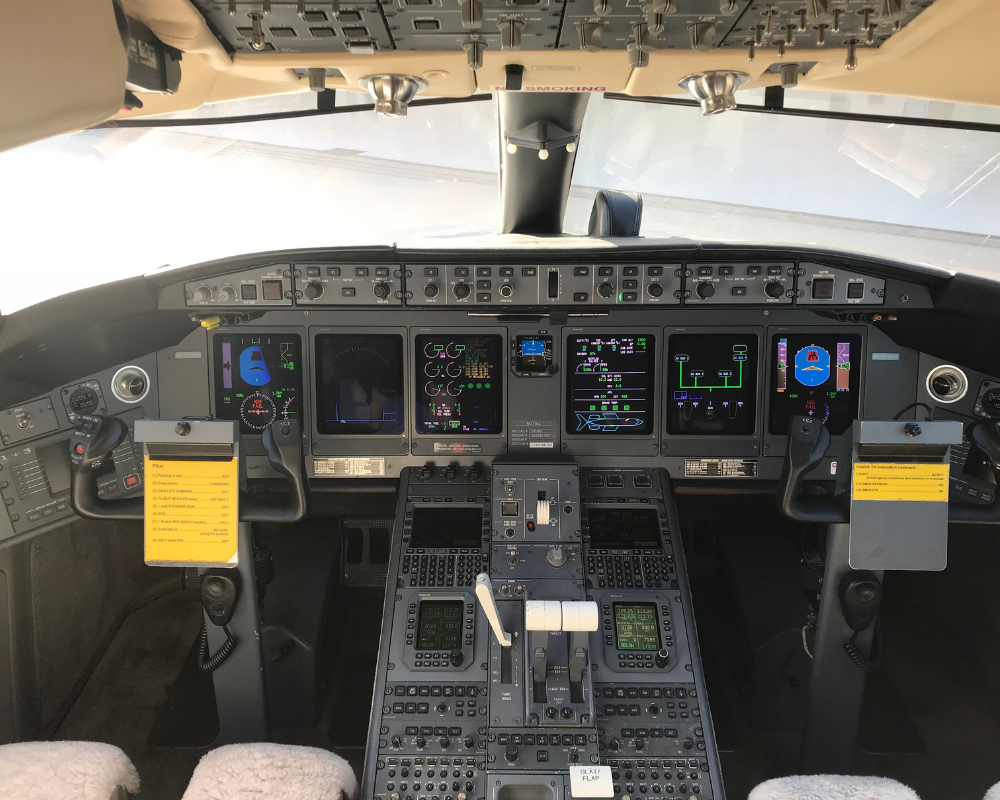 Cockpit of a jet aircraft, showing instrument panels, screens, controls, and pilot's view through the windshield.