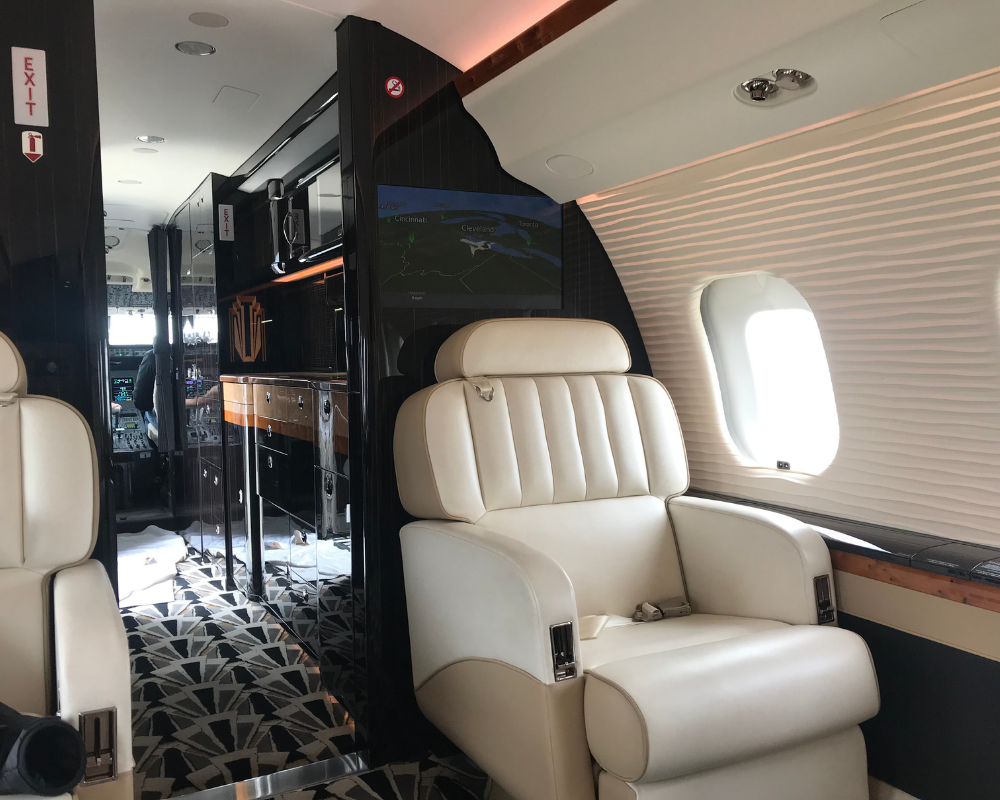 Interior of a private jet cabin, featuring beige leather seating, black and wood paneling, and patterned flooring.