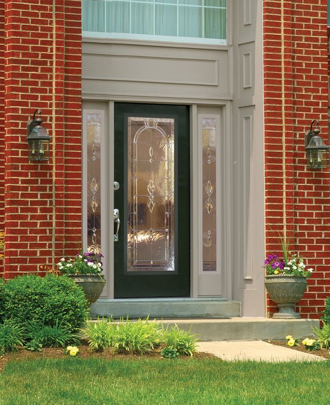 Brick house front door with glass panel, flanked by lanterns, flower pots, and neatly trimmed shrubs.