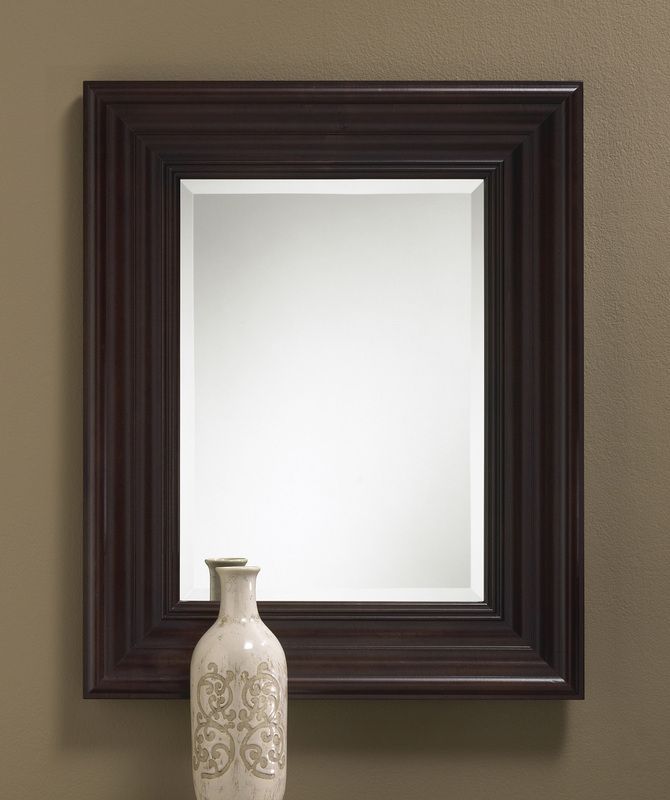 Dark-framed wall mirror above a white decorative vase on a brown wall.