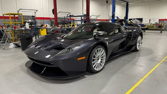 Gray sports car in a garage, parked on a yellow-lined floor beside workshop equipment.