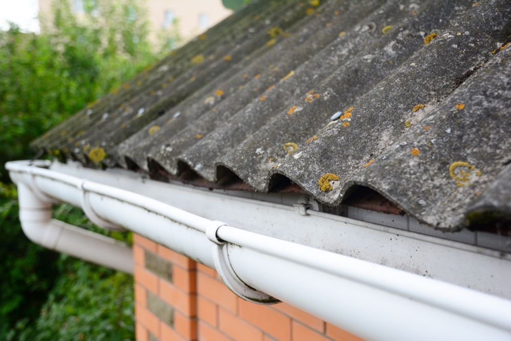 Close-Up of An Asbestos Roof with A White Gutter Along a Brick Building — Geoff Hall Roofing in Frederickton, NSW