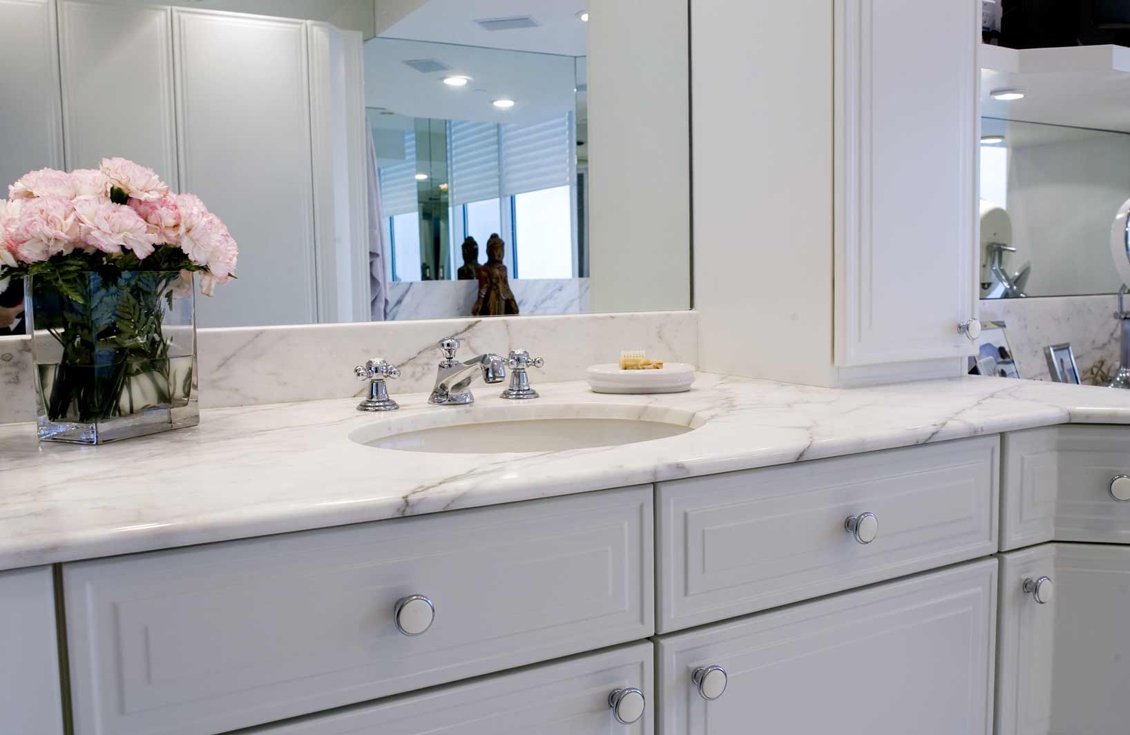 A bathroom with white cabinets , a sink , a mirror and a vase of flowers