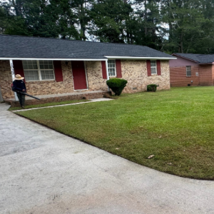 A person using a leaf blower on the lawn in front of a brick house on a residential street.