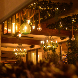 A low-angle view of a restaurant patio at night, illuminated by warm hanging lights, green glass globes, and lush greenery.