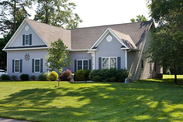 A grey, two-story suburban house with a dark shingled roof, set behind a lush green lawn on a sunny day.