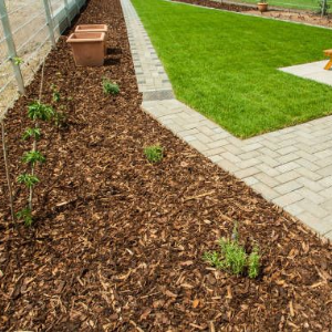 A backyard with a brown mulch garden bed, paved walkway, green grass, and a garden hose reel on a patio.