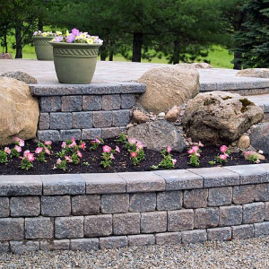 A stone retaining wall with tiered levels and built-in steps, featuring pink flowers in a bed and a decorative planter.