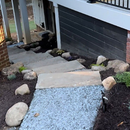 A stone walkway leads to concrete steps under a raised wooden deck with black railings and a black dog resting near them.