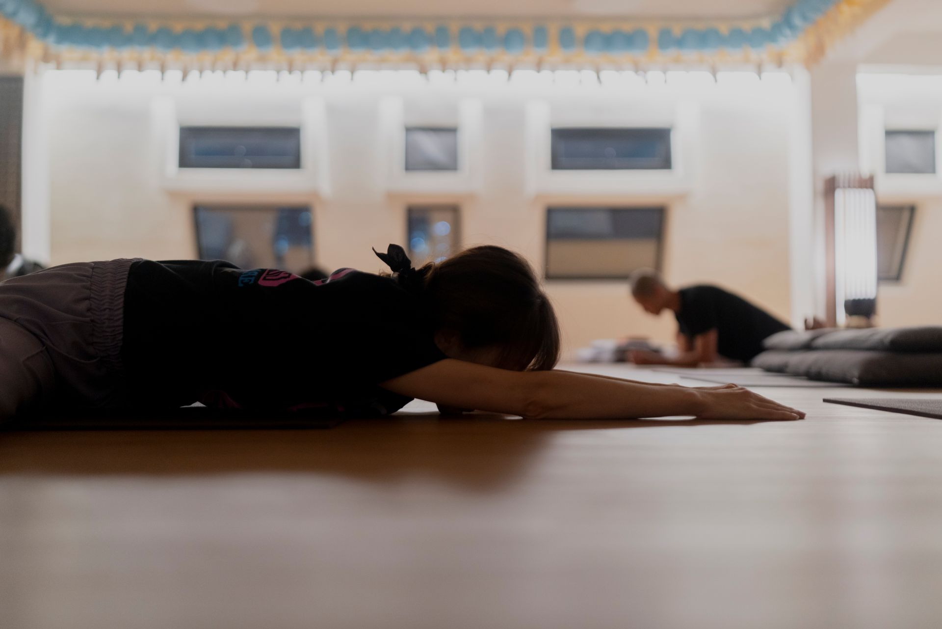 Woman in yoga pose, chest to floor, arms extended; others in background, indoor setting.