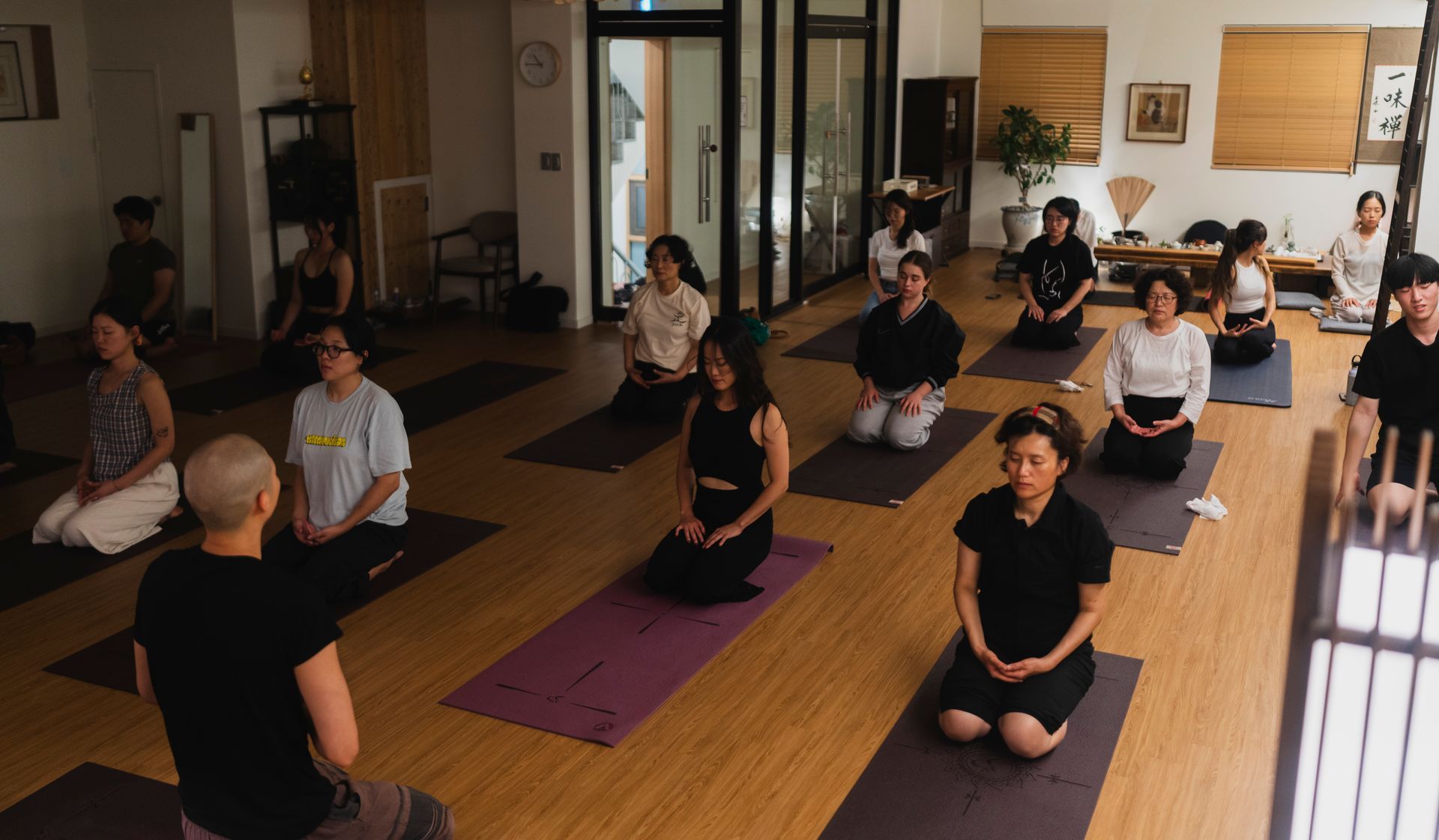 People seated in meditation pose on mats in a bright room with a teacher facing them.
