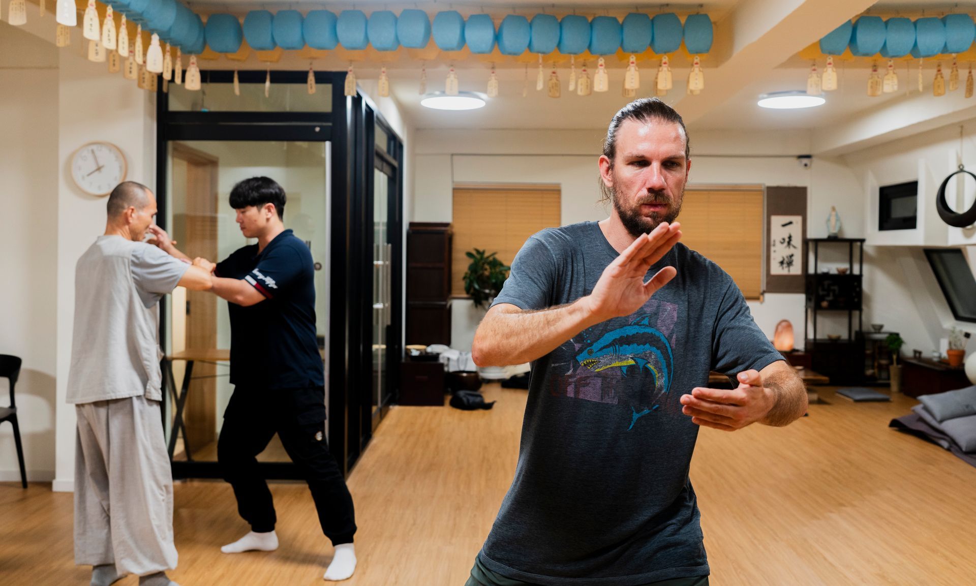 Man practicing Tai Chi in a studio, two other people in background, light wood floor.