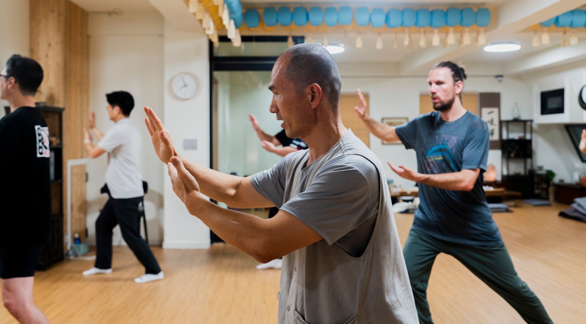 People practicing Tai Chi in a studio, with arms extended. One man in focus.