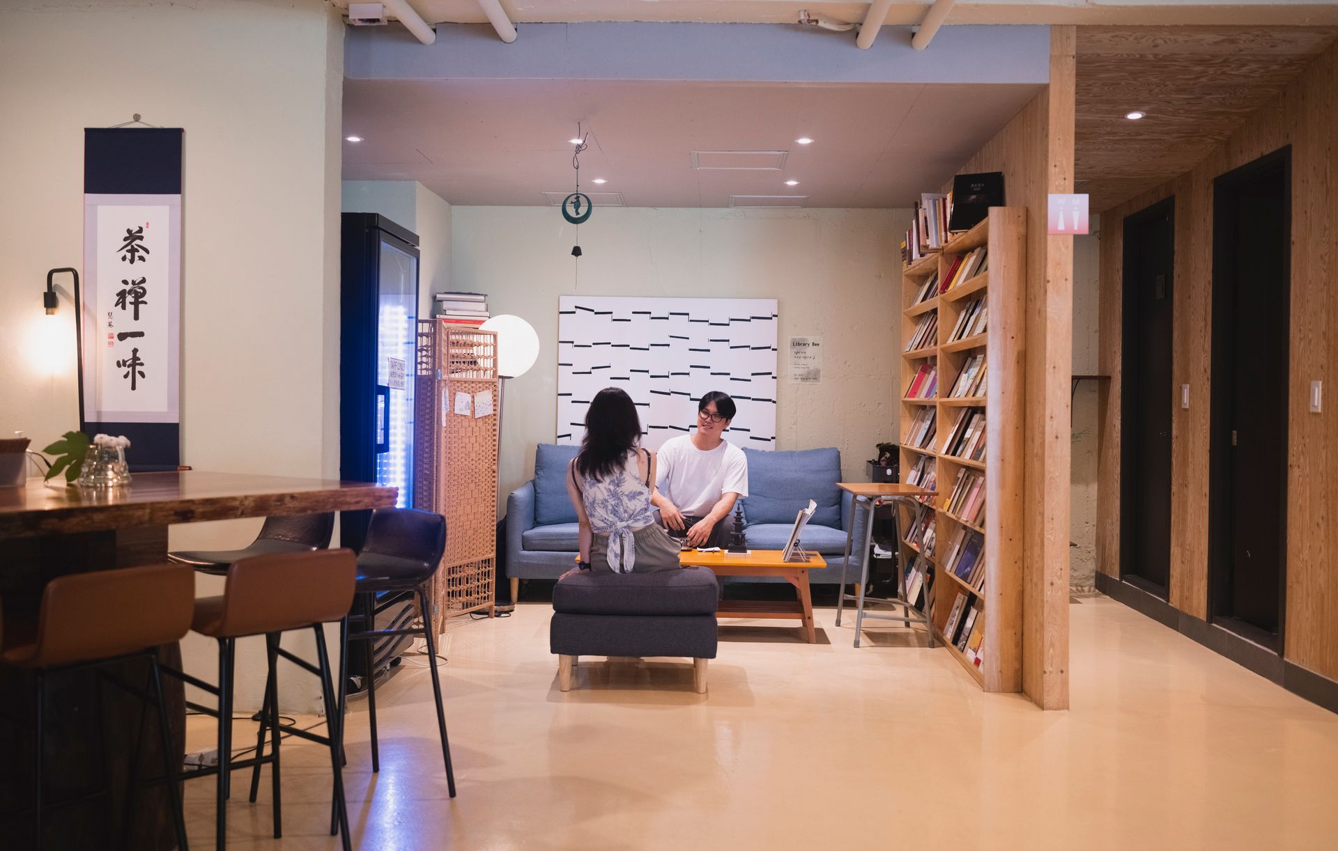 Couple in a living room with a bookshelf. The woman and man are seated. Beige and wood tones.
