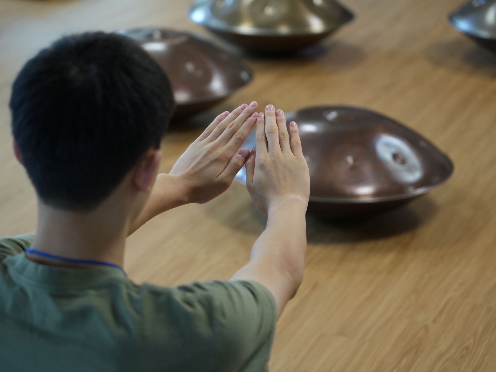 Person facing handpans, hands raised, possibly playing. Brown instruments on wooden floor.