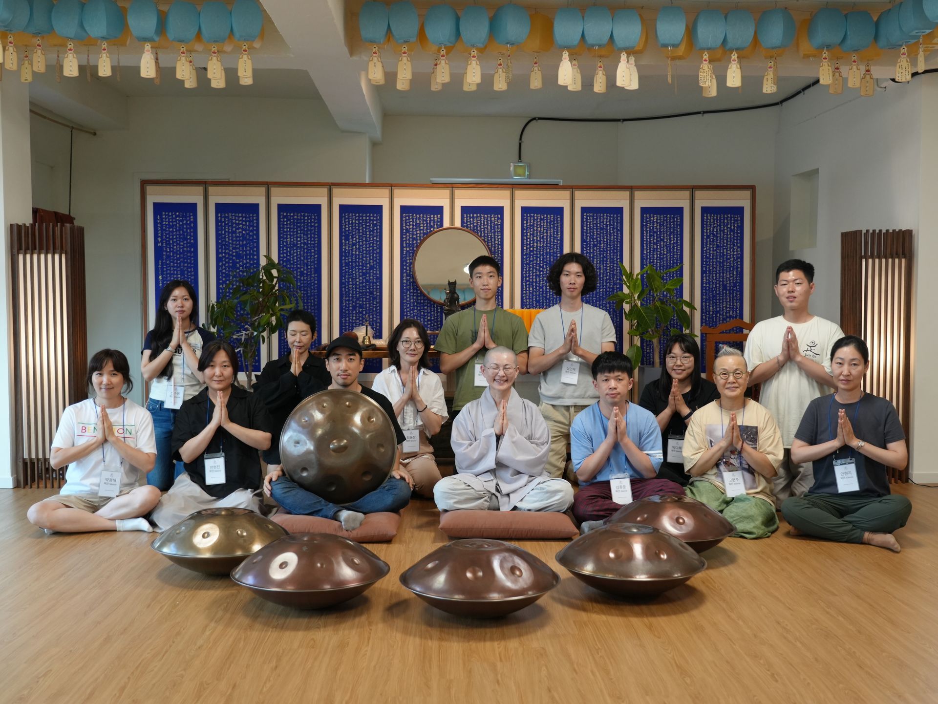 Group of people with hands in prayer pose, gathered with handpans, in a room with traditional decor.