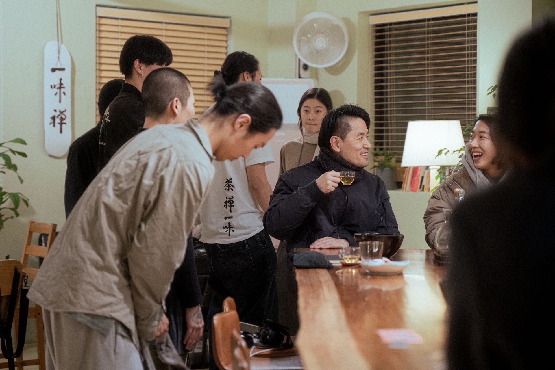 People gather at a bar. A man in a coat sips tea, smiling. Others stand or sit, some laughing.