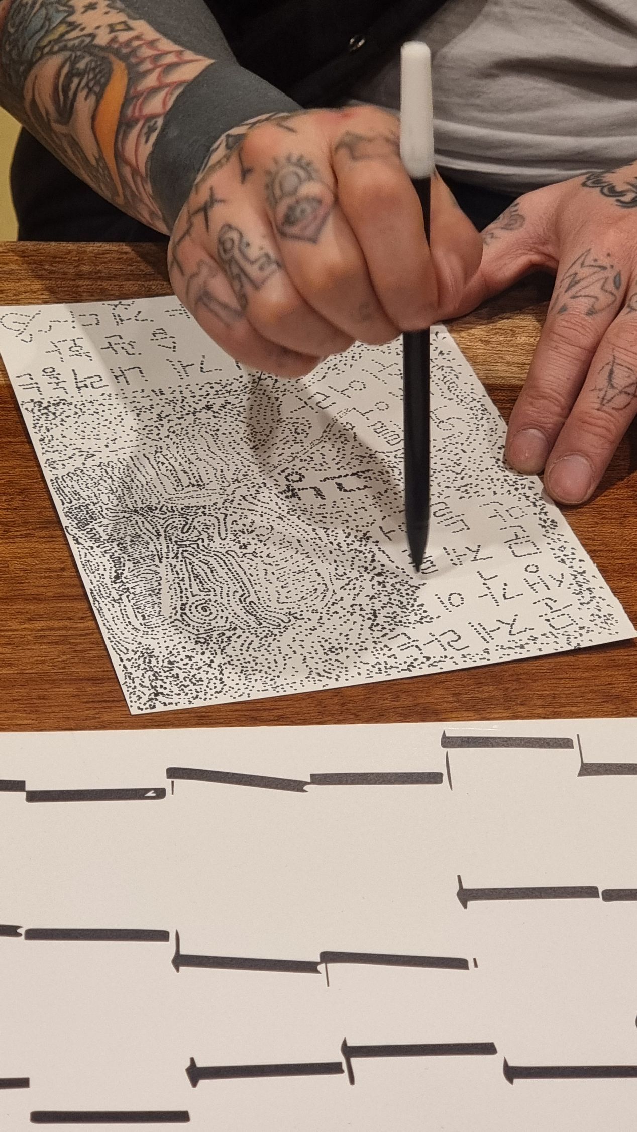Person with tattooed arms drawing black lines on paper with a pen.