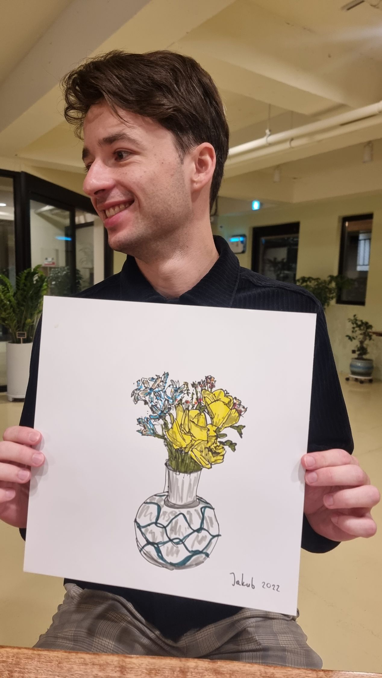 Man smiling, holding a drawing of flowers in a vase, indoors.