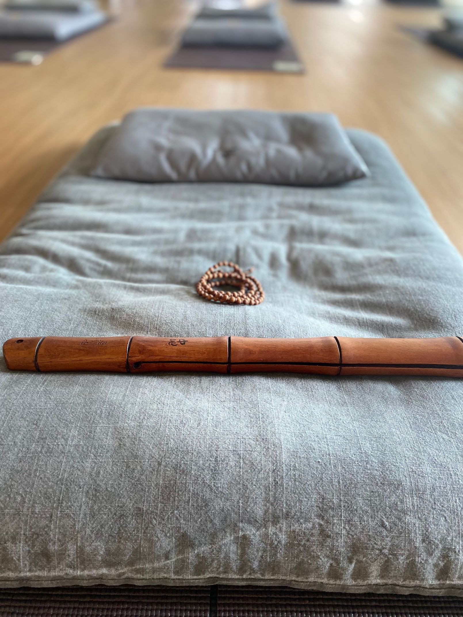 A meditation mat with a wooden flute, beads, and a pillow on a wooden floor.