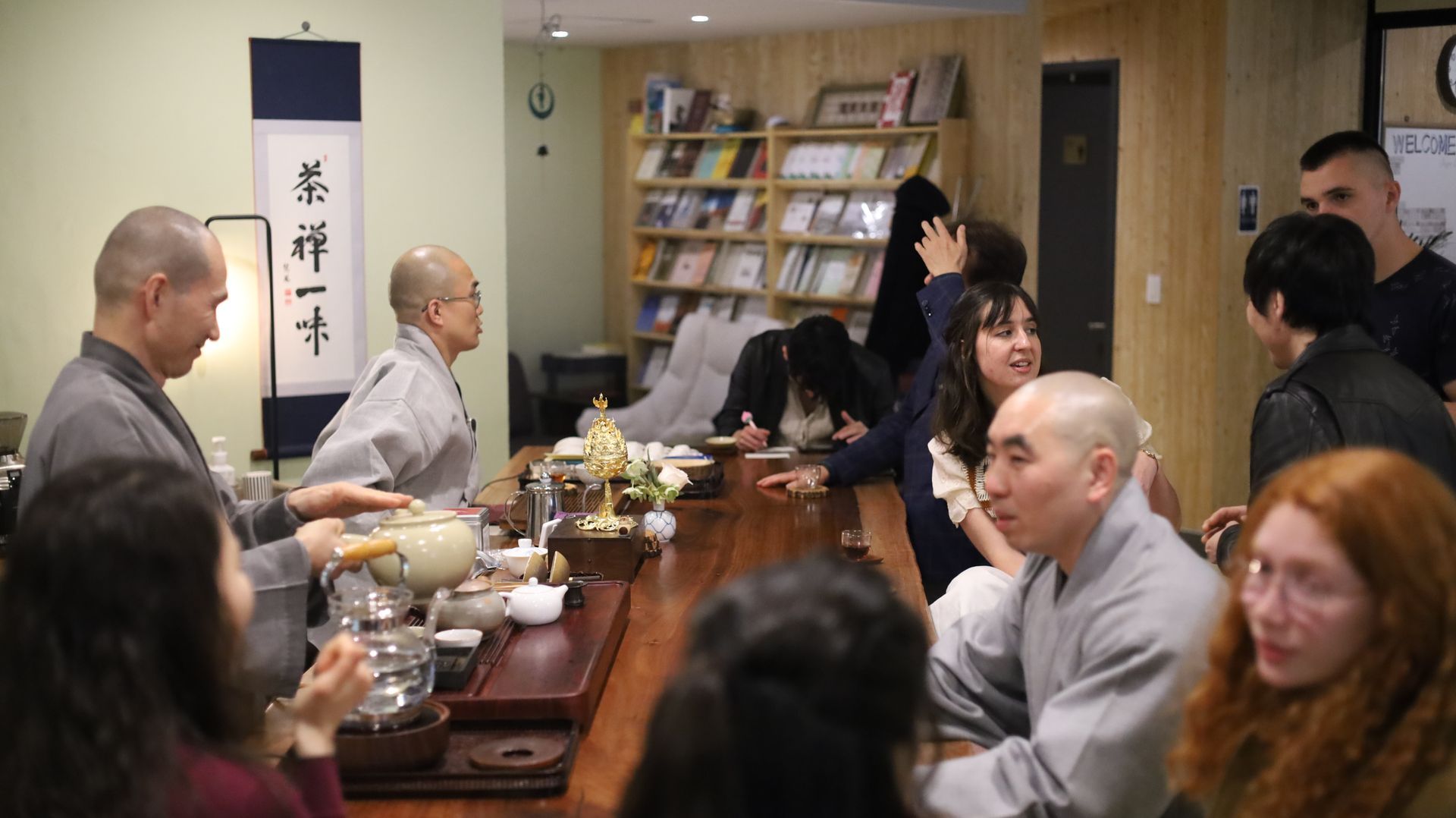 사람들은 긴 탁자, 불교 승려, 책이 있는 방에 모여 차를 마십니다.