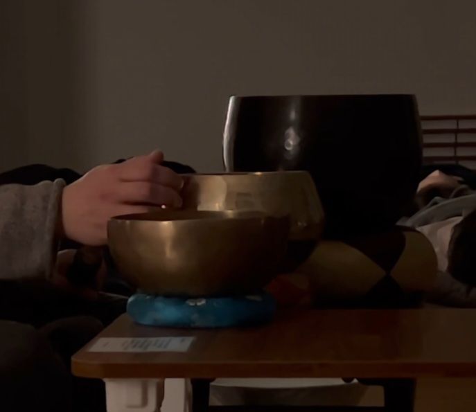 A person’s hands near two bronze singing bowls on a table; low light setting.