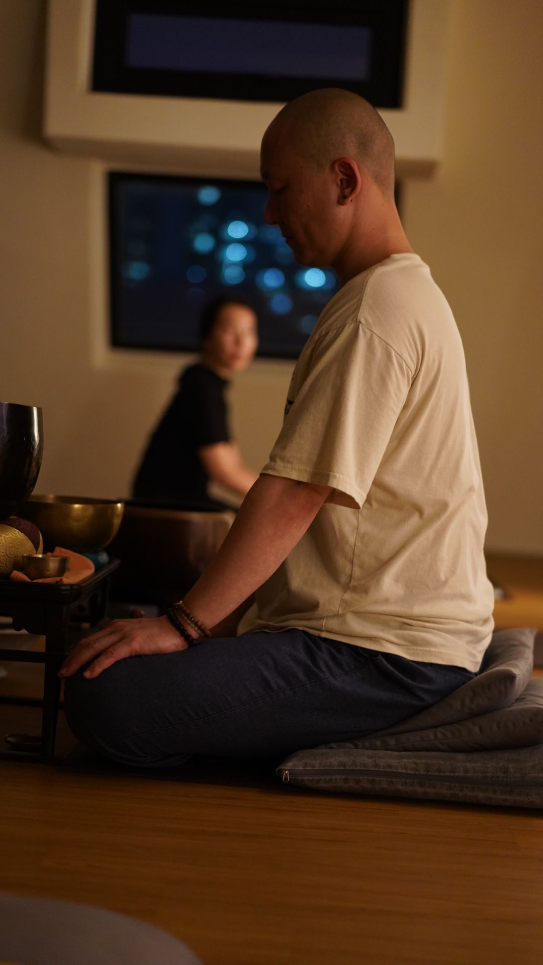 Man with shaved head meditating indoors, with another person in the background; warm lighting.