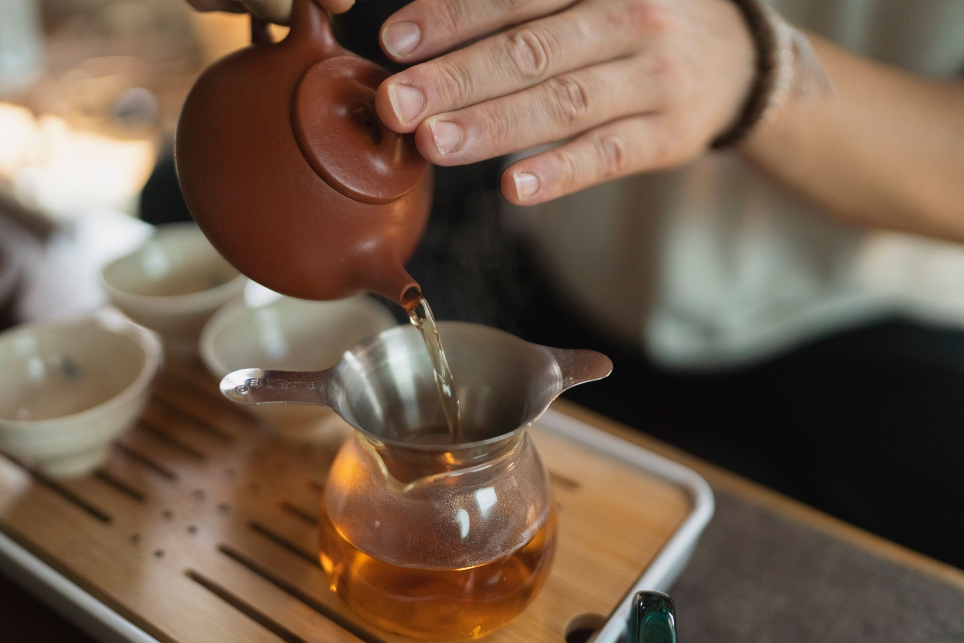 Person pouring tea from a brown teapot into a glass pitcher, through a strainer. Teacups and wooden tray are in view.
