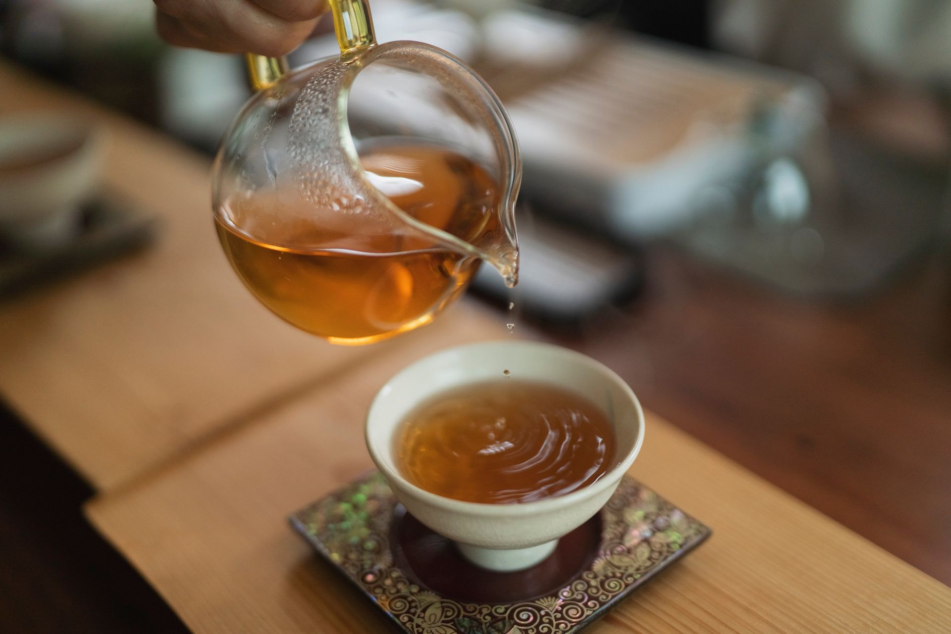 Person pouring tea from a glass pitcher into a white teacup, close up. Wooden surface, warm tones.