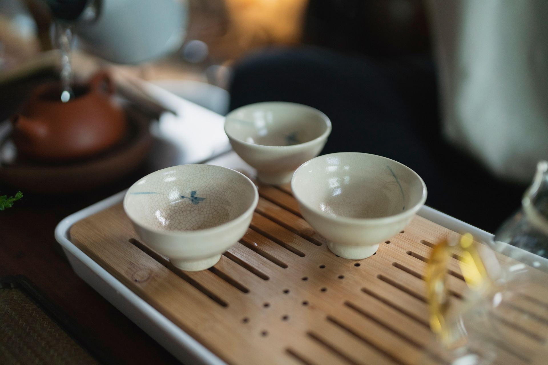 Three small, beige tea cups on a wooden tray. Tea is being poured from a brown teapot.